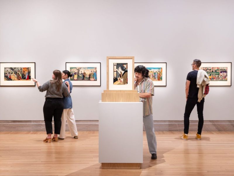 一群人在观看白色画廊墙上展示的装裱好的艺术品，前景是白色基座上的中央展品。