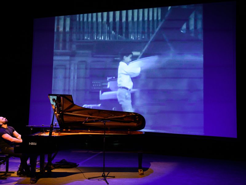 A musician plays a grand piano on stage, while behind him, a large black-and-white image of a person swinging an axe is projected on a screen—a true showcase of Adam Tendler's flair for experimental music.