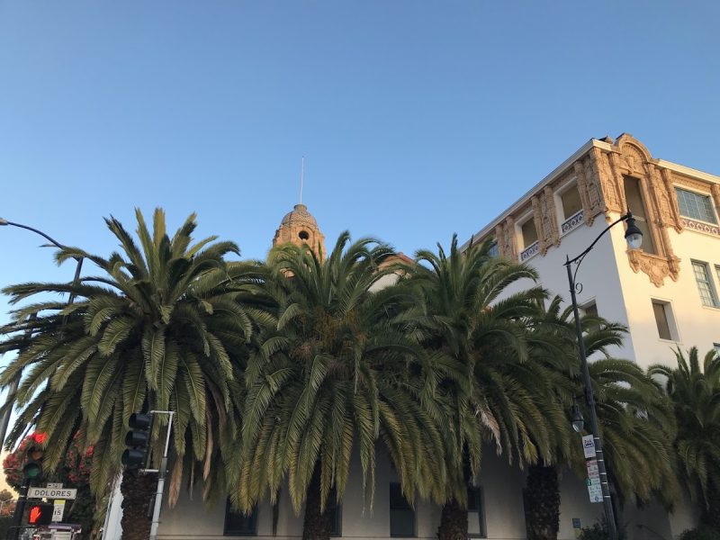 A white building with ornate architectural details is partially obscured by tall palm trees under a clear blue sky.