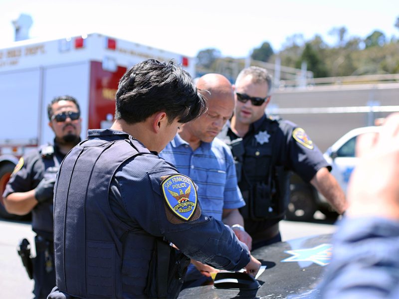 Police officers in uniforms are standing around a man in a blue shirt near a police vehicle, inspecting documents on the car's hood. Another emergency vehicle is in the background, suggesting the man may be homeless and in need of assistance.