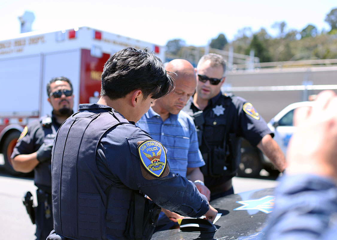 Police officers in uniforms are standing around a man in a blue shirt near a police vehicle, inspecting documents on the car's hood. Another emergency vehicle is in the background, suggesting the man may be homeless and in need of assistance.