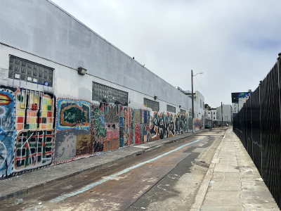 A long alleyway with colorful graffiti on the walls of a white industrial building on the left and a black fence on the right. The sky is overcast.