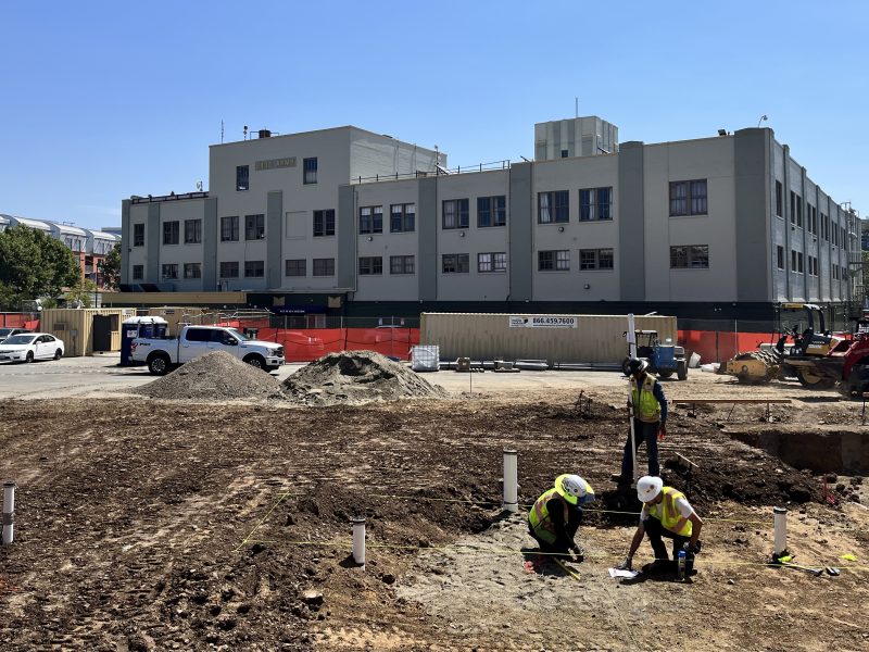 Three construction workers in safety vests and hard hats work on a cleared construction site with pipes. A large building is visible in the background, and a construction truck is parked nearby.