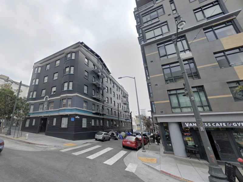 Urban street corner featuring apartment buildings, a crosswalk, parked vehicles, and a nearby café with visible signage.