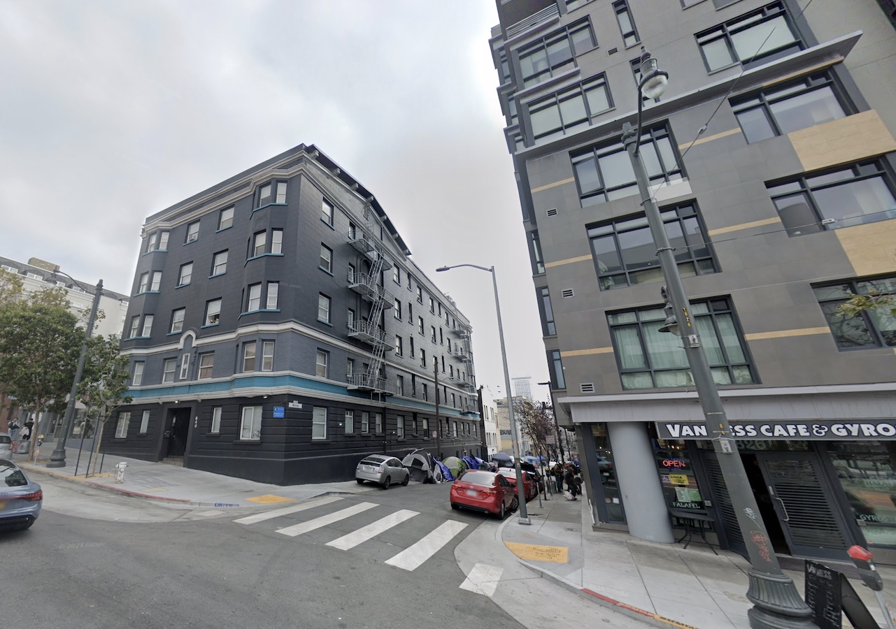 Urban street corner featuring apartment buildings, a crosswalk, parked vehicles, and a nearby café with visible signage.