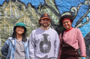 Three people standing in front of a graffiti-covered wall, all smiling. One person wears a green hat, another wears a baseball cap, and the third wears a red headband.