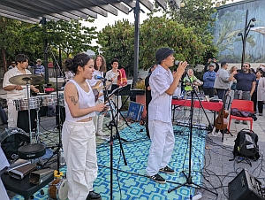 A band performs outdoors on a patterned mat with two vocalists singing into microphones. Drummer and other musicians play in the background. An audience watches the performance.