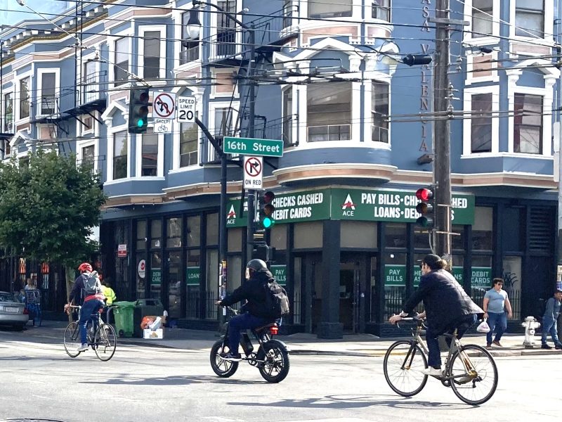 Three cyclists ride down a street in an urban area with buildings and a store offering financial services in the background. The intersection is marked with traffic signs and signals.