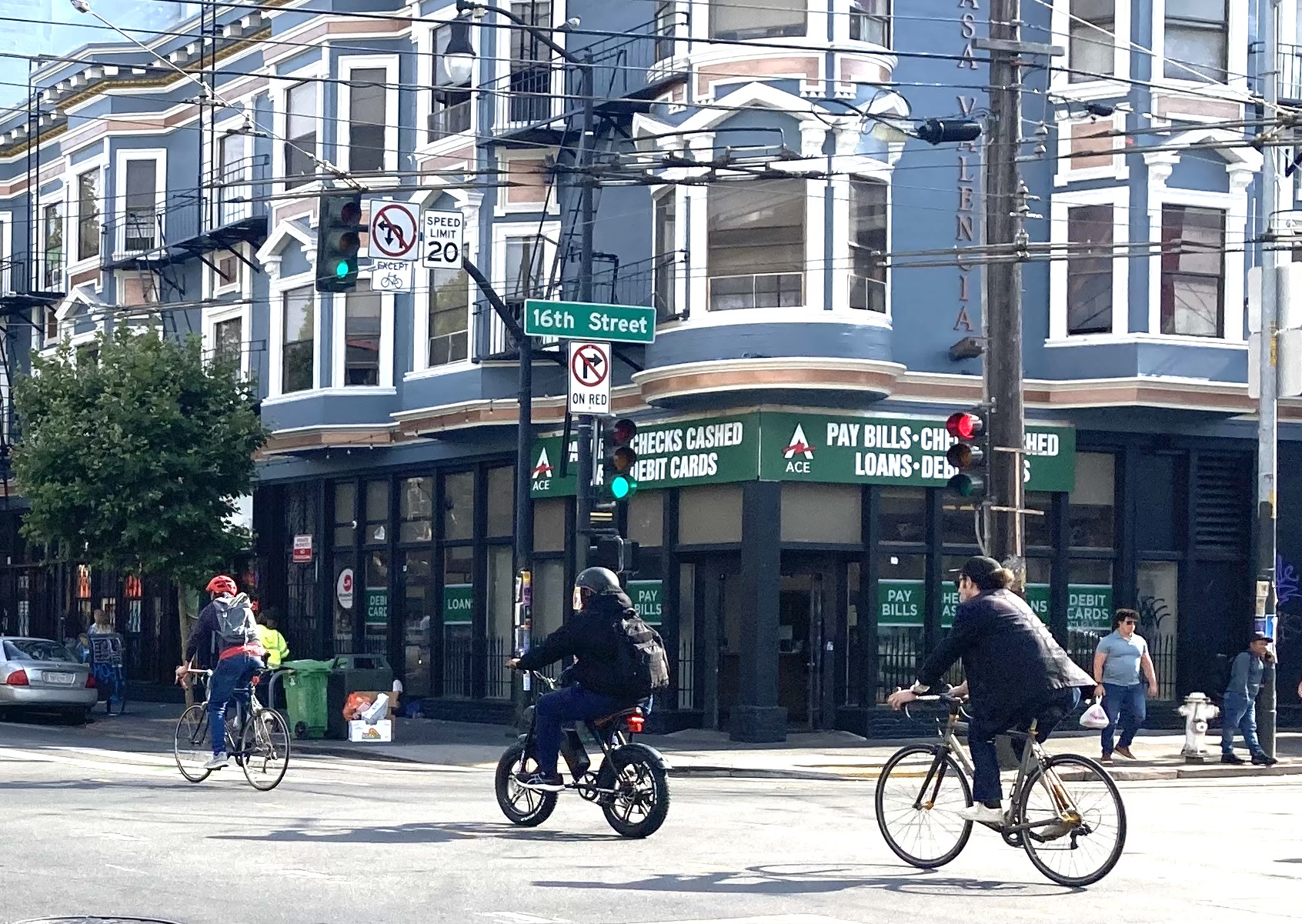Three cyclists ride down a street in an urban area with buildings and a store offering financial services in the background. The intersection is marked with traffic signs and signals.