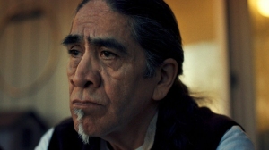 A man with long black hair and a goatee looks pensively into the distance.