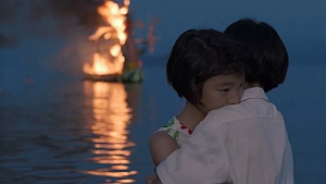A child is embraced by an adult near a body of water at dusk, while a boat is burning in the distance on the water.