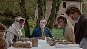 A group of people, dressed in rustic costumes with face paint, sit around a table outside near a stone building. They all look towards the camera with a serious expression.