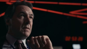 A man in a suit and tie looks pensively to the side, illuminated by red and dark lighting in the background.
