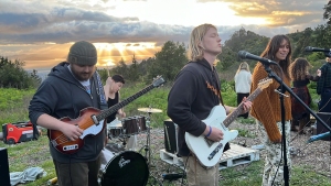 A band performs outdoors at sunset. The group includes a guitarist, a drummer, another guitarist, and a vocalist. Several people are in the background. Trees and a partially cloudy sky are visible.