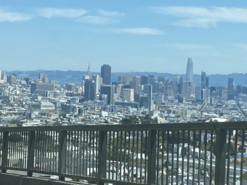 A clear daytime view of a city skyline with a variety of tall buildings and a bridge railing in the foreground.
