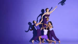 Five dancers in purple activewear perform an artistic, elevated pose against a purple background. The central dancer balances on the others, reaching upwards while their shadows cast on the wall.
