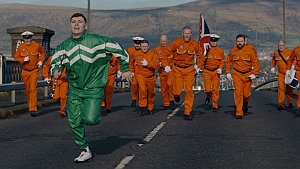 A person in a green tracksuit is running on a road, followed by a group of people in orange uniforms holding a flag.