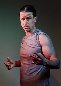 A person in a sleeveless gray shirt poses with hands slightly raised, looking directly at the camera.