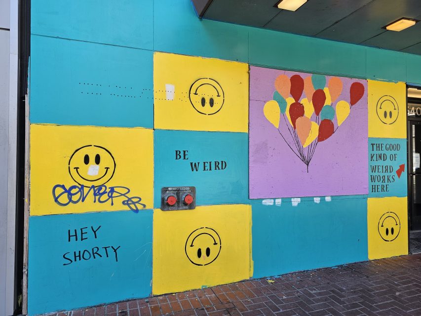 A wall painted with yellow smiley faces, the phrases "BE WEIRD" and "HEY SHORTY," graffiti, and a colorful mural of balloons with the text "THE GOOD KIND OF WEIRD WORKS HERE.