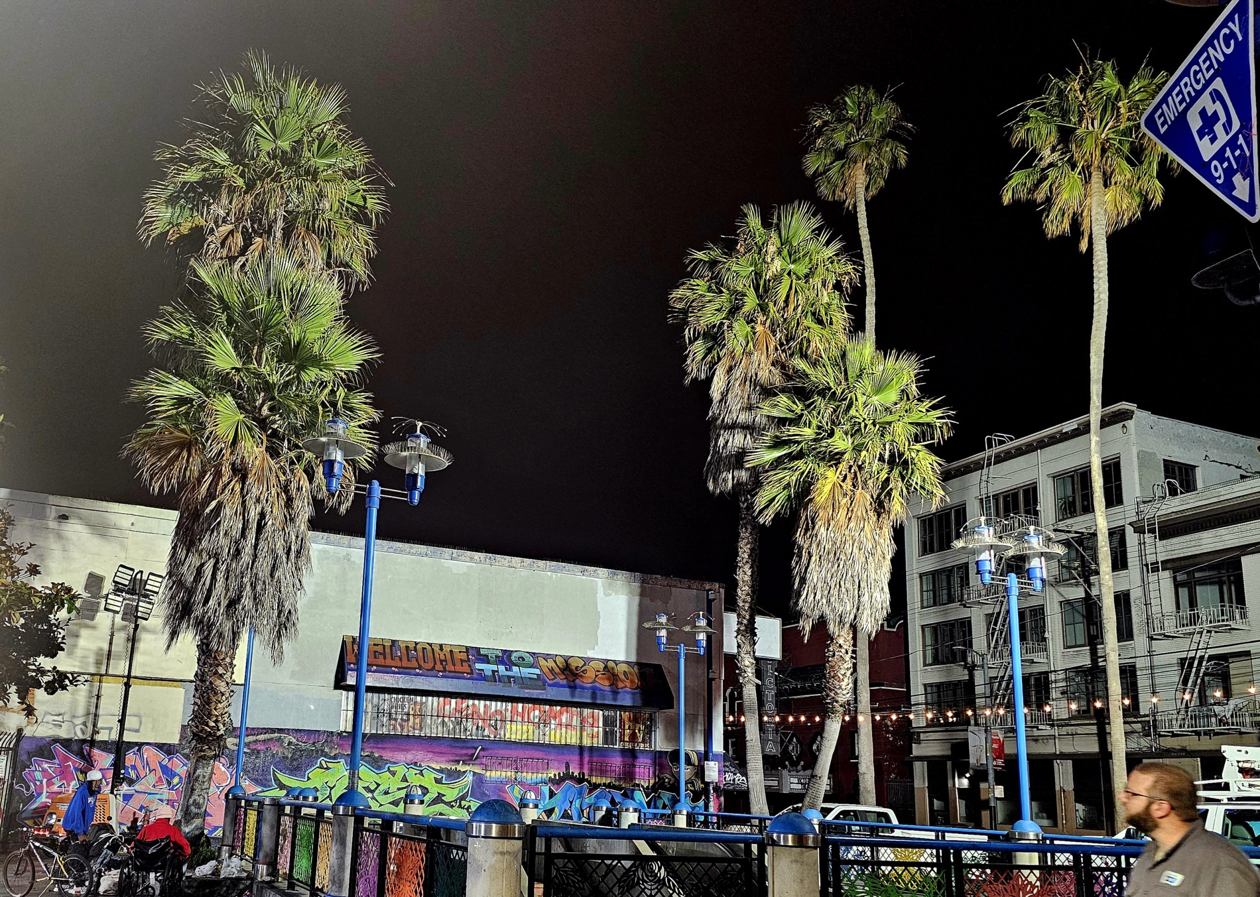 A nighttime scene with palm trees, graffitied walls, and string lights. People walk through the area, and a blue emergency sign is prominently displayed. Buildings are visible in the background.
