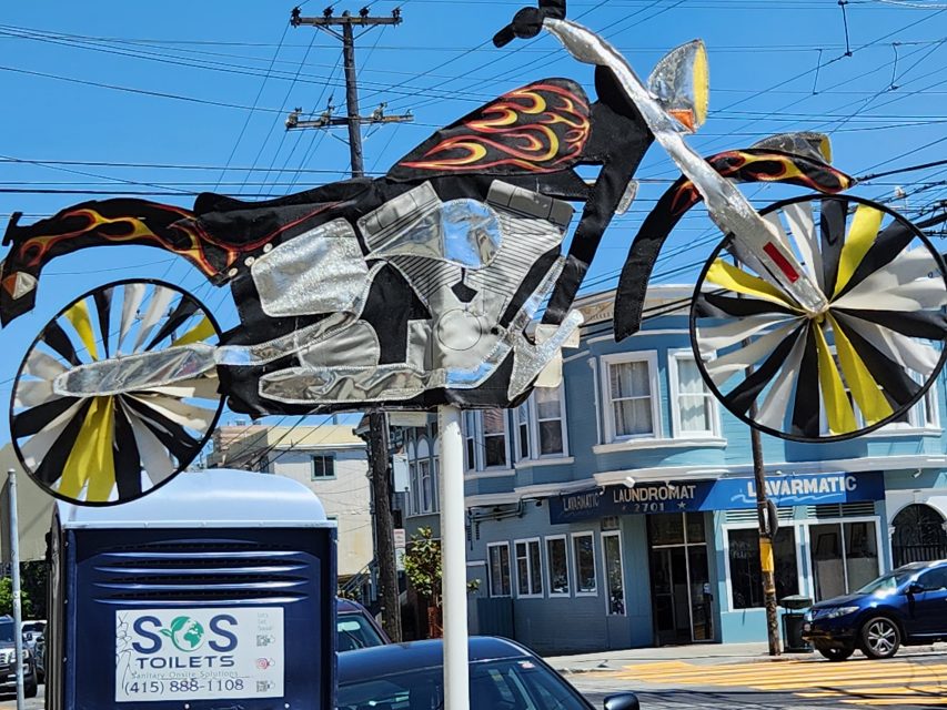 Metal sculpture of a motorcycle with flame designs stands on a pole near a portable toilet with signs in front of buildings and power lines.