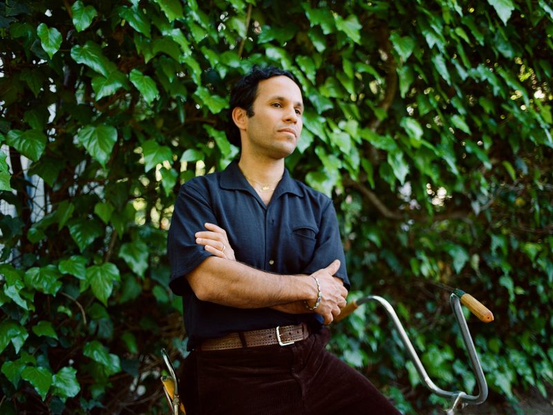 a young Puerto Rican-American man stands with arms crossed in front of a wall of ivy. He's wearing a blue shirt and black pants