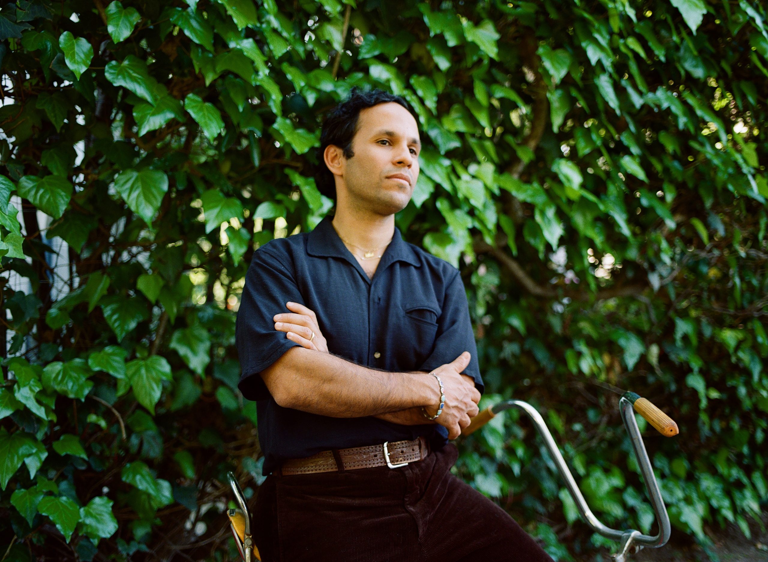 a young Puerto Rican-American man stands with arms crossed in front of a wall of ivy. He's wearing a blue shirt and black pants