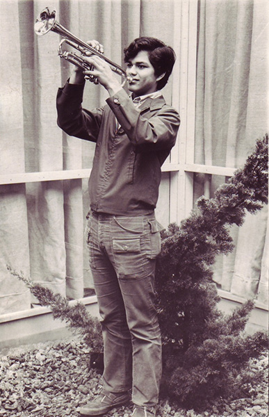 Black-and-white photo of a young man standing and playing a trumpet outside, with a small tree and curtains in the background.