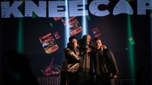 Three individuals stand closely together on stage with the word "KNEECAP" displayed prominently behind them. The background features colorful, floating images.