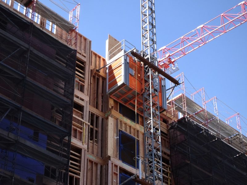 A multi-story housing building under construction, with scaffolding and a red crane. An orange construction elevator is attached to the right side of the structure. The sky is clear and blue.