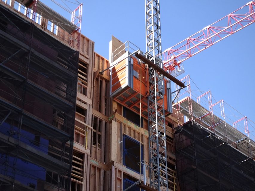 A multi-story housing building under construction, with scaffolding and a red crane. An orange construction elevator is attached to the right side of the structure. The sky is clear and blue.