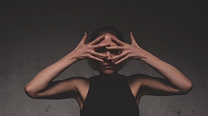A person with a dark background holds their hands in front of their face, fingers splayed.