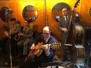 A quartet of musicians in suits performs in a dimly lit room with yellow walls. One plays the saxophone, another the guitar, a third the double bass, and the fourth, in the background, plays the trumpet.