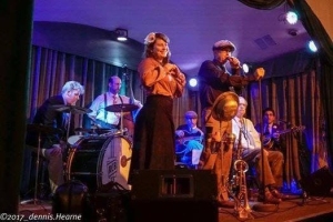 A band performs on stage with a female singer and a male musician in the foreground. Other band members play various instruments, including drums, a guitar, and a trumpet. Stage lights illuminate the scene.