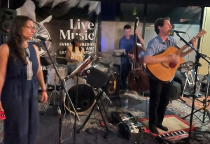 Musicians perform live on stage with a guitarist, vocalist, drummer, bassist, and keyboard players. A "Live Music" sign is visible in the background.