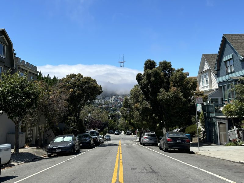A street in a hilly residential area with parked cars and houses on both sides. A fog-covered hill and a tall, red and white tower are visible in the background.