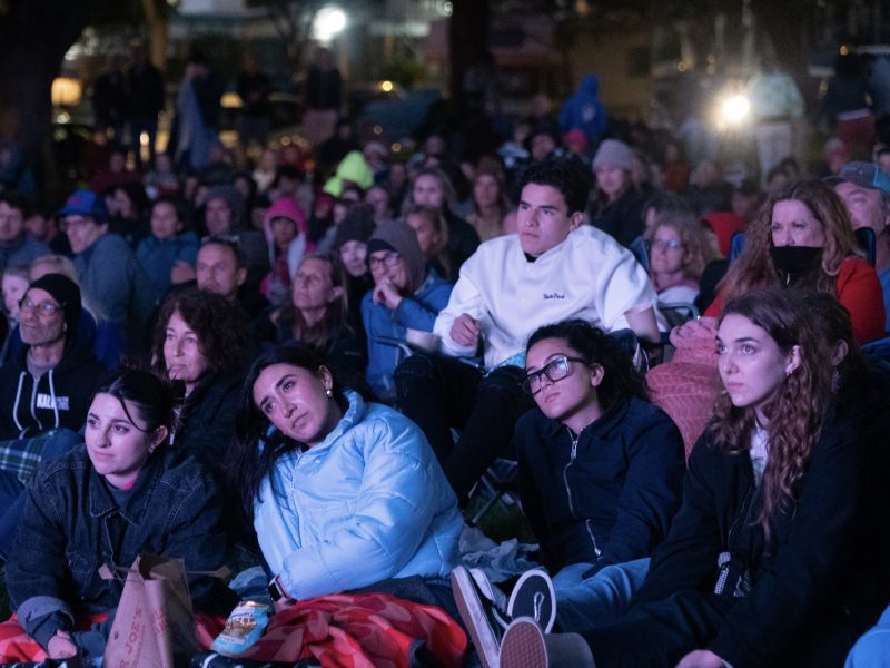 A large group of people sit closely together on the ground, outdoors at night, watching an event. Most are wearing jackets and some have blankets, indicating chilly weather.