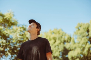 A man wearing a black t-shirt and cap looks upwards while standing outdoors with a background of green trees and a clear blue sky.