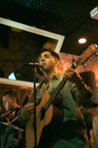 A man plays an acoustic guitar and sings into a microphone on stage, while another musician plays an accordion in the background.