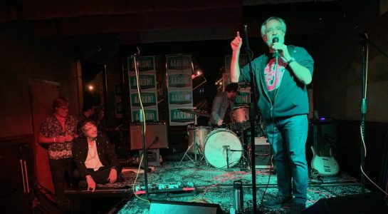 A man singing into a microphone on a stage with other people.