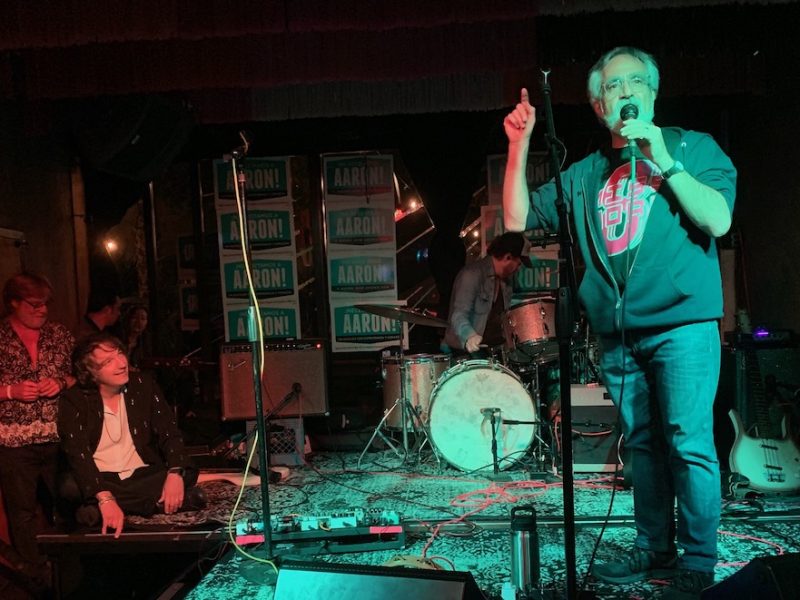 A man singing into a microphone on a stage with other people.