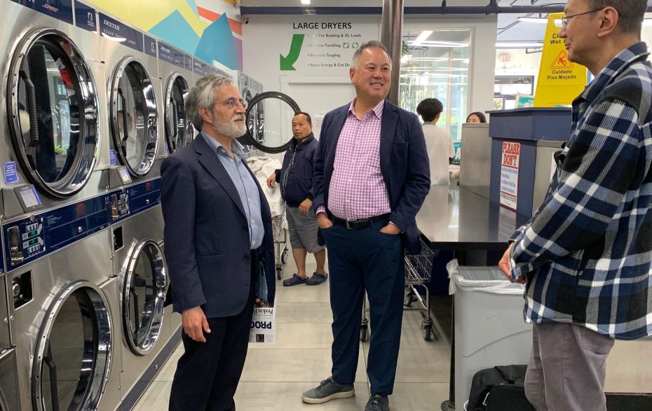 Three men are standing and conversing inside a laundromat, with large dryers visible in the background. Two men are in formal attire, while one is dressed casually in a plaid shirt.