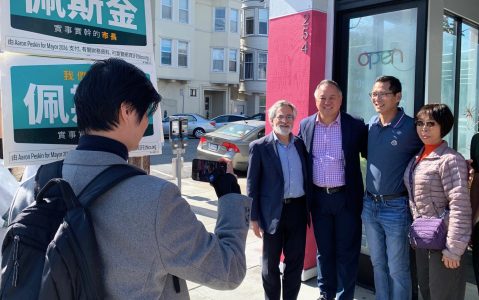 Four people are posing for a photo outside a building, with a man taking their picture. The building door has the word "open" on it, and there are signs with text in both English and Chinese in the background.