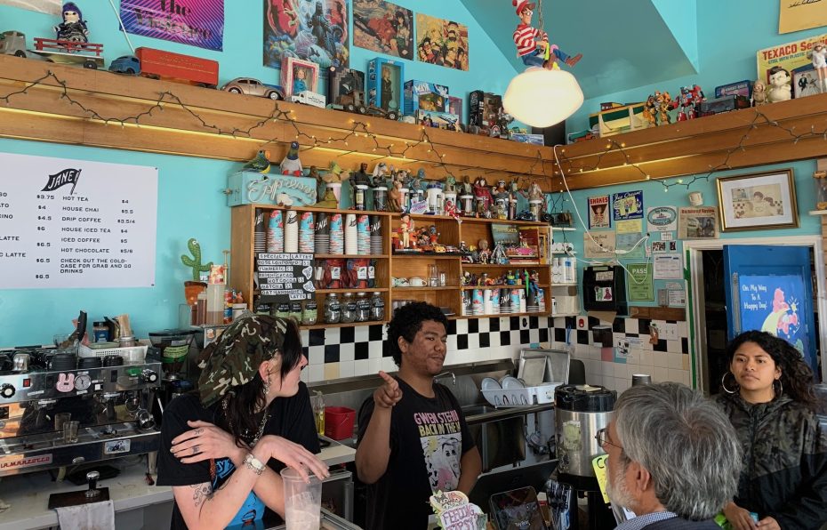 A small, brightly colored café with three baristas attending to customers. The counter is decorated with various toys and posters, and a menu with drink options is visible on the wall.