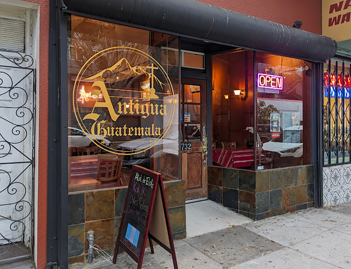 Exterior of a restaurant named "Antigua Guatemala" with a large circular logo on the glass window and a chalkboard menu outside. The entrance door has a "732" number, and a lit "OPEN" sign is visible.