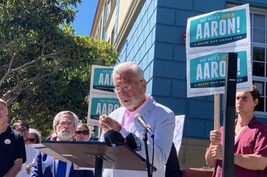 A man in a white jacket speaks at a podium with microphones, surrounded by people holding signs that read "We need Aaron! Mayor who knows how.