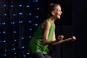 A woman wearing a green sleeveless top and gray pants is performing on stage with string lights in the background. She is smiling and has her hands raised in expressive gestures.