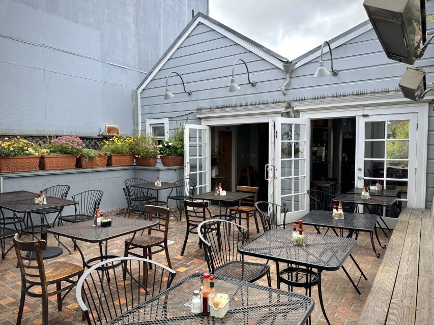 Outdoor patio with black metal tables and chairs, potted plants, and an open door leading inside. Bottles of condiments and napkins are on the tables. Overcast sky in the background.