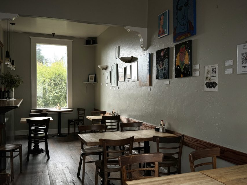 An empty cafe with wooden tables and chairs, a large window letting in natural light, and various framed artworks displayed on the walls.