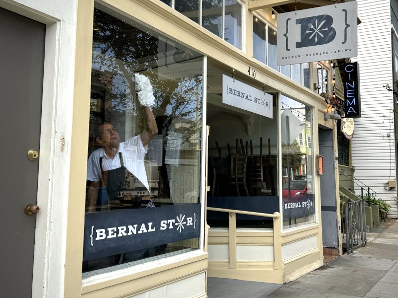 A person is cleaning the front window of Bernal Star restaurant. The reflection of trees and a building is visible in the glass. There is a sign for a cinema on the adjacent building.